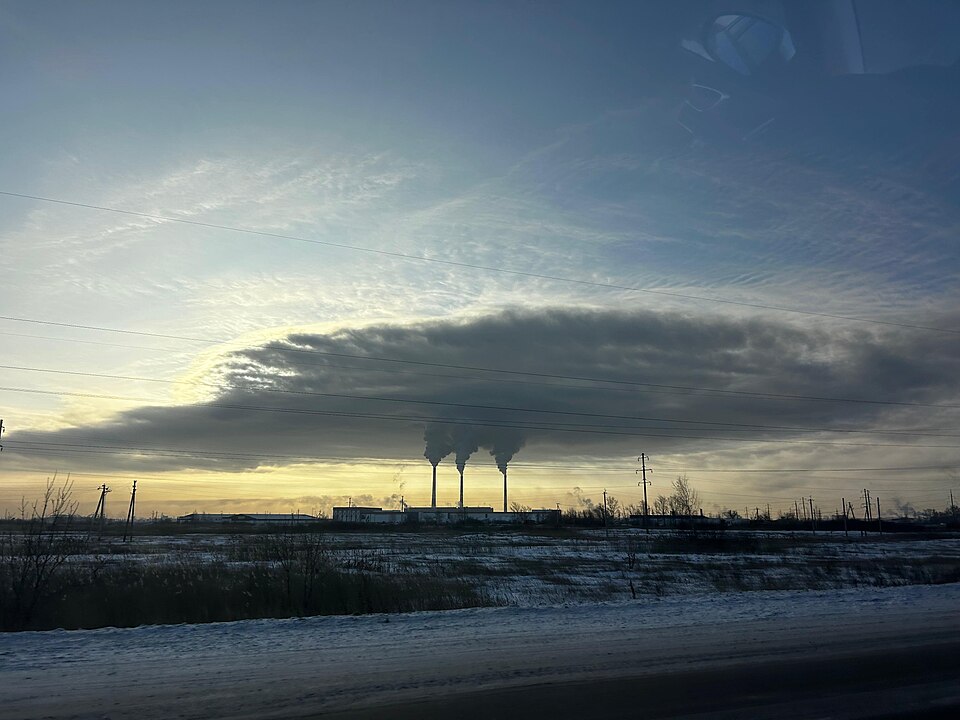 A coal-fired power station on the horizon at dusk, its three chimneys trailing a long dark plume of flue gas across the sky