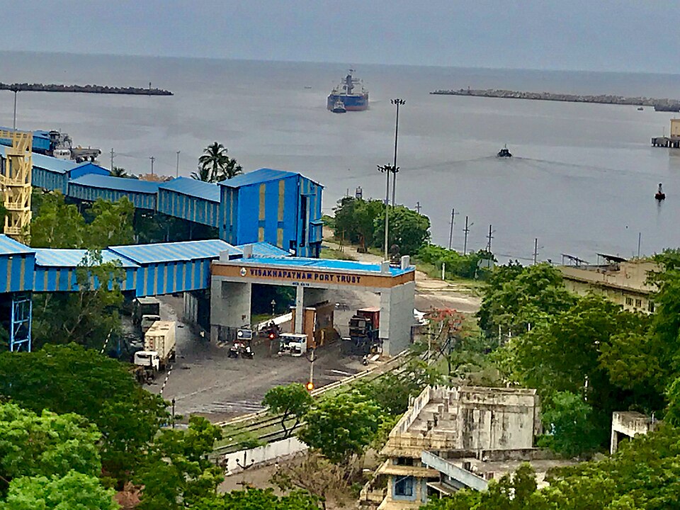 Visakhapatnam Port Trust gate below a hill, with the sheltered harbour basin behind it, a cargo ship at the entrance and breakwaters on either side of the approach channel