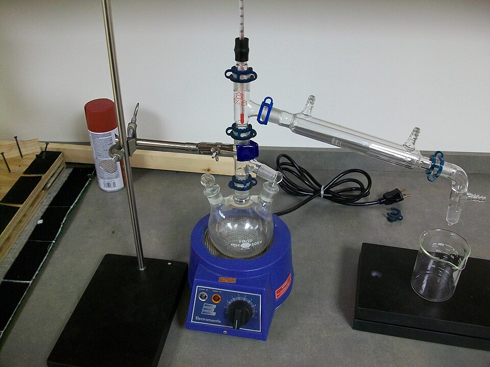 Laboratory distillation set-up: a round-bottom flask on an electric heating mantle, a thermometer in the still head and an inclined water condenser leading to a receiving beaker