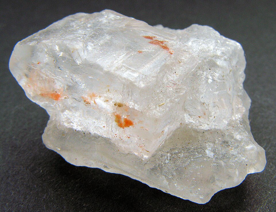 A colourless glassy halite crystal showing stepped cubic cleavage faces, with small orange mineral inclusions, on a dark background