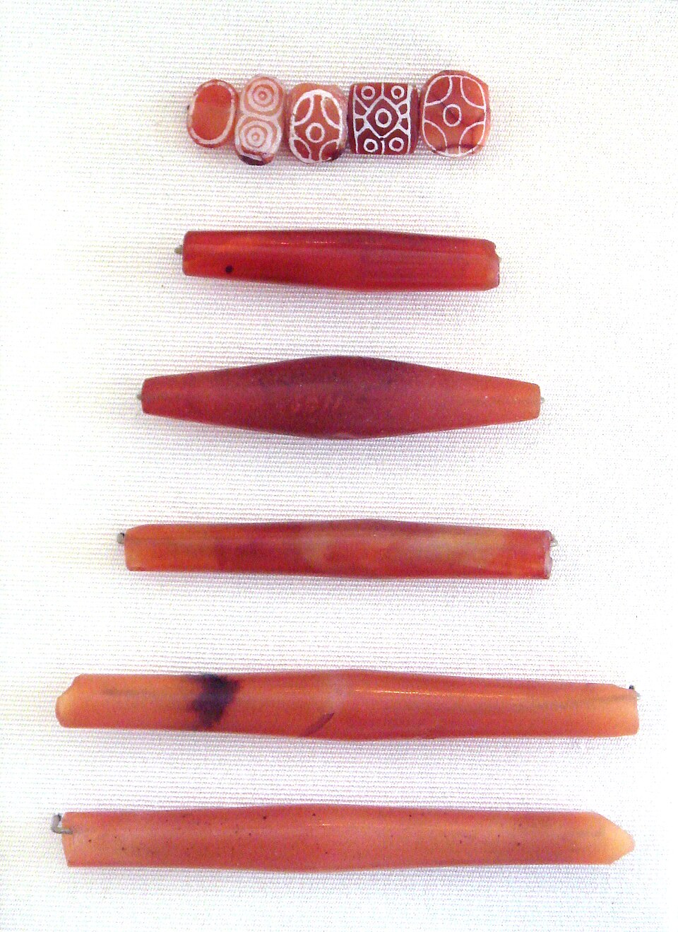 Indus Valley carnelian beads laid on white cloth: five short beads etched with white circle and eye patterns above four long tapering barrel beads