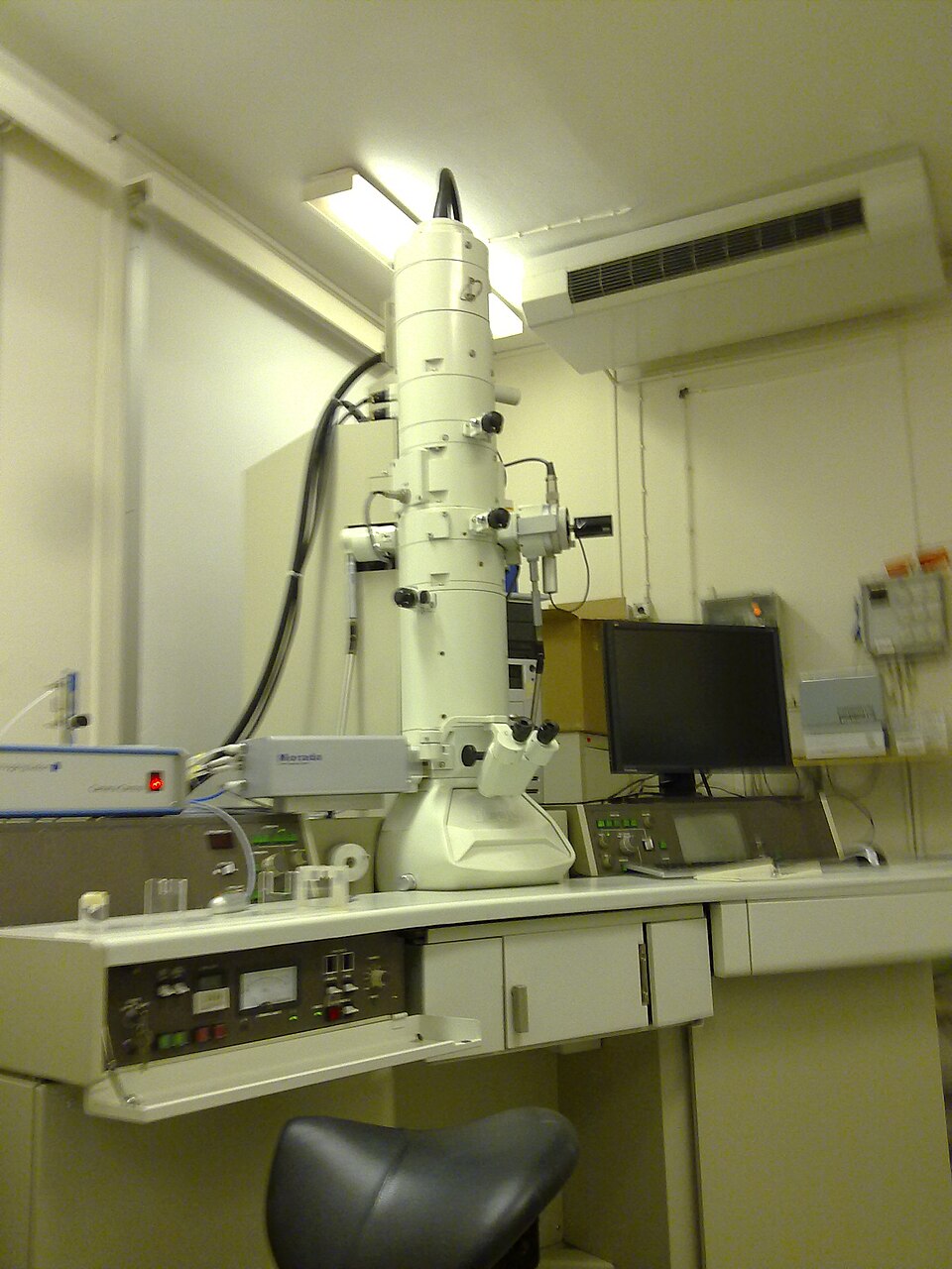 A transmission electron microscope in a laboratory: a tall white vertical column with a binocular viewing port, standing on a control console beside a monitor