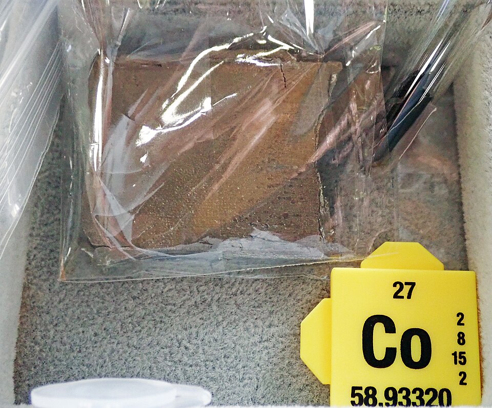 A bagged slab of grey-brown cobalt metal beside a yellow element card reading Co, 27, 58.93320