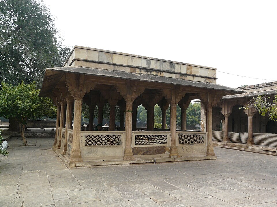 The tomb of Tansen at Gwalior: a low open pavilion of carved sandstone pillars with pierced stone screens, standing on a paved plinth beside a tamarind tree