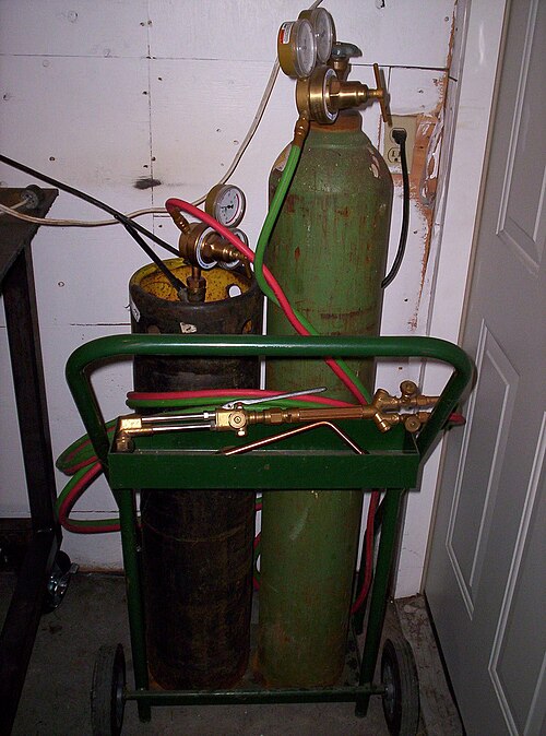 An oxy-acetylene welding set: acetylene and oxygen cylinders on a green trolley, with regulators, red and green hoses and a brass torch
