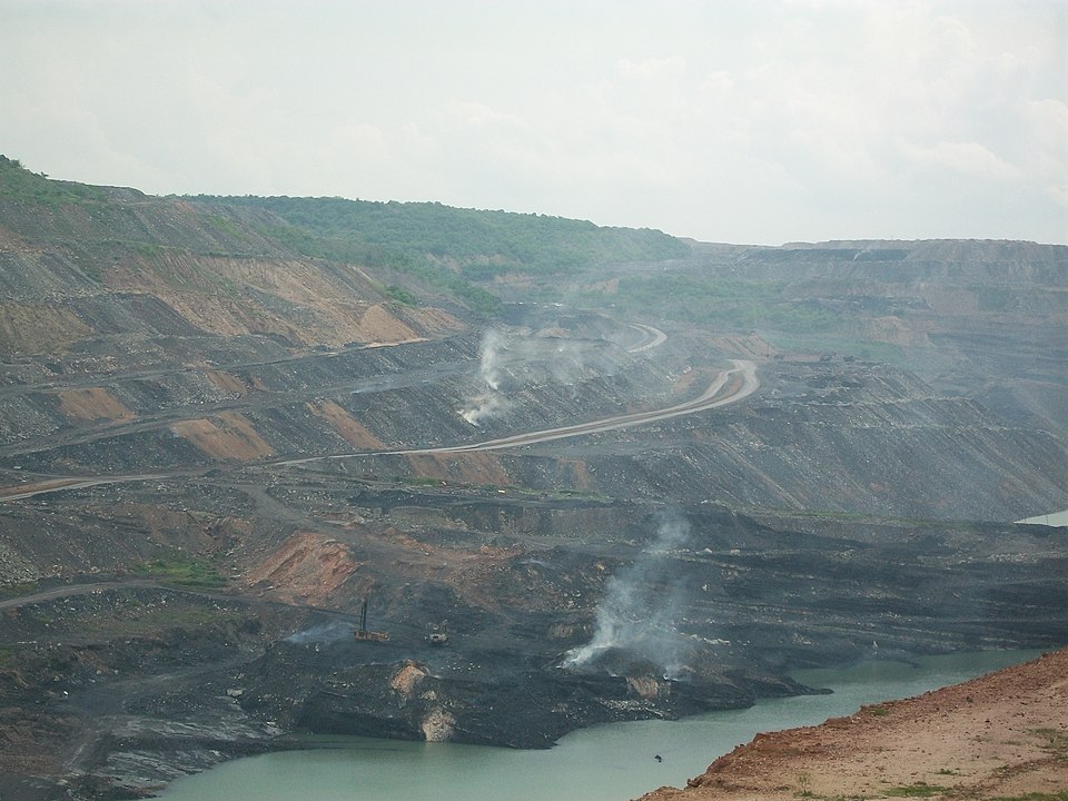 A wide opencast coal mine cut in stepped black benches, with haul roads winding between them and water pooled in the pit floor