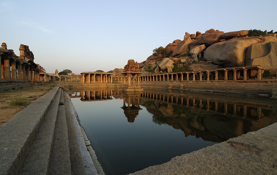 A long stepped stone tank at Hampi reflecting the colonnades along its banks, with a small pillared shrine at the centre and granite boulder hills behind