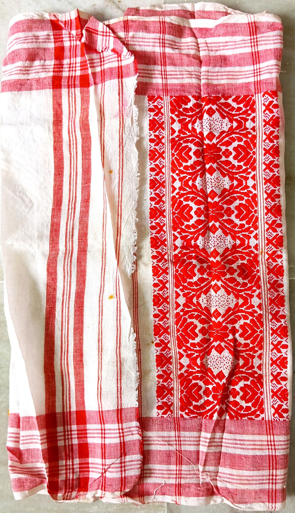 A white handwoven cotton gamosa laid flat, edged with red stripes and carrying a broad band of woven red floral motifs at one end