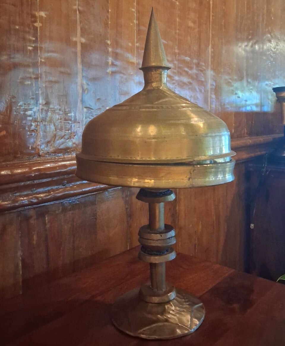 An Assamese xorai in bell metal standing on a wooden table: a tray on a turned pedestal foot, closed with a domed cover topped by a tall spike