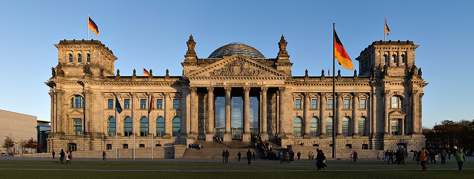 The west front of the Reichstag building in Berlin at sunset, its portico inscribed DEM DEUTSCHEN VOLKE with the glass dome rising behind and German flags flying