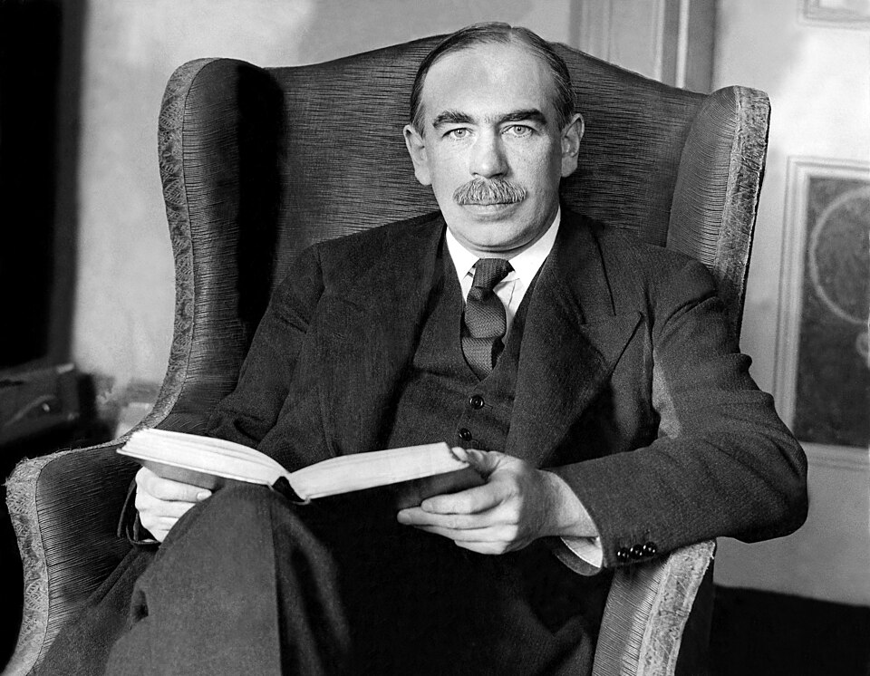 Black-and-white photograph of John Maynard Keynes seated in a wing armchair in a three-piece suit, holding an open book