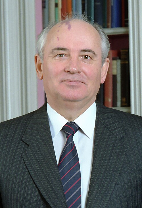 Colour portrait of Mikhail Gorbachev in a dark pinstriped suit and striped tie, standing in front of a bookshelf