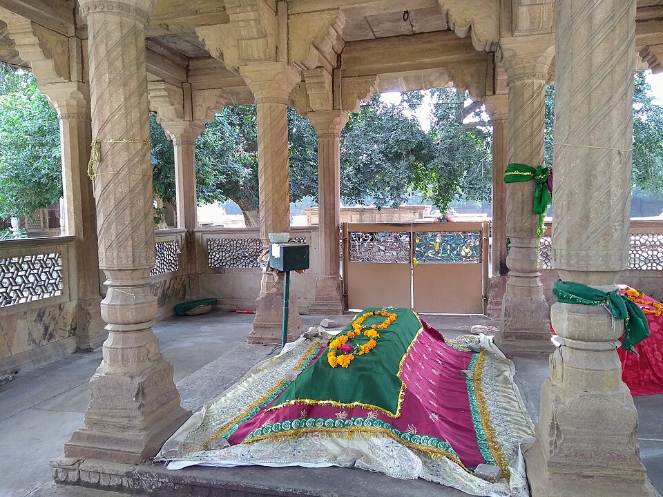 Tansen's grave at Gwalior under an open sandstone pavilion, the tomb draped in green and pink cloth with marigold garlands, a tamarind tree visible beyond the pillars