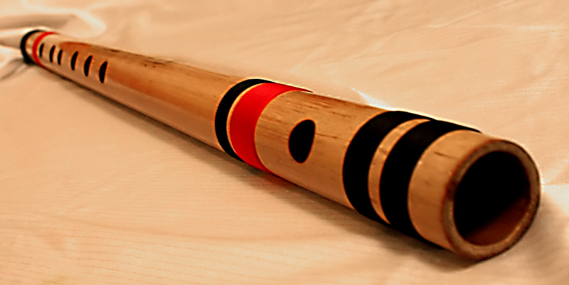 A bamboo bansuri lying on a cloth, its side-blown embouchure hole and row of finger holes visible along the tube between red and black thread bindings.