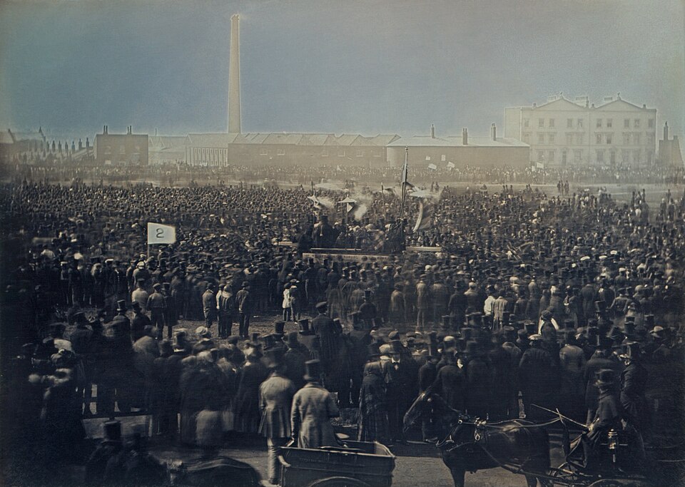 An 1848 daguerreotype of a vast crowd in top hats packed onto Kennington Common around a speakers' platform and banners, with London factory chimneys behind.