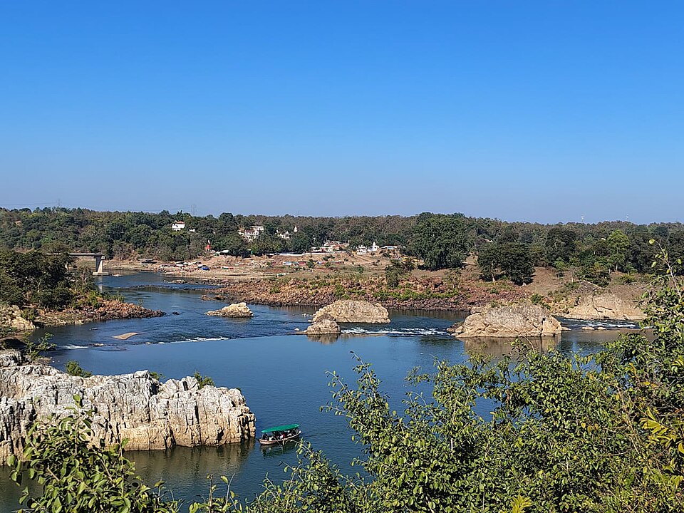 The Narmada River winding past white marble rock ledges at Bhedaghat near Jabalpur, with a small boat on the water and wooded banks beyond.