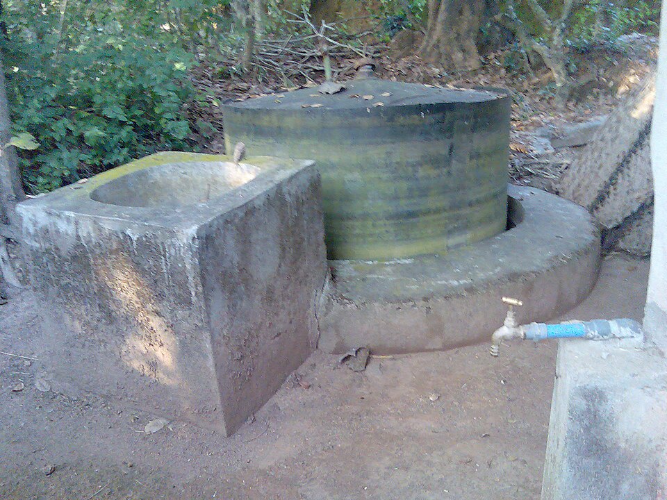 A village gobar gas plant: a concrete inlet mixing tank beside a sealed cylindrical gas holder drum resting in a water seal, with an outlet tap.