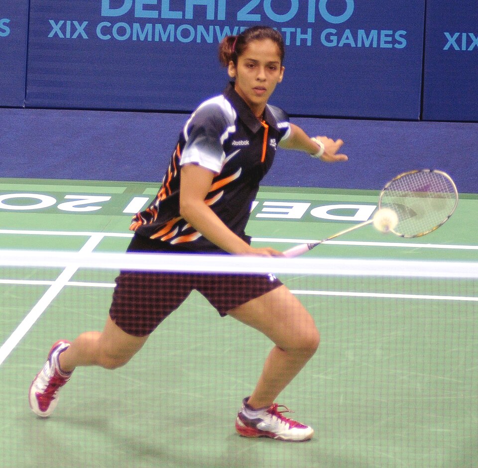 Saina Nehwal lunging forward to play a forehand shot at the net during a badminton match on a green court.