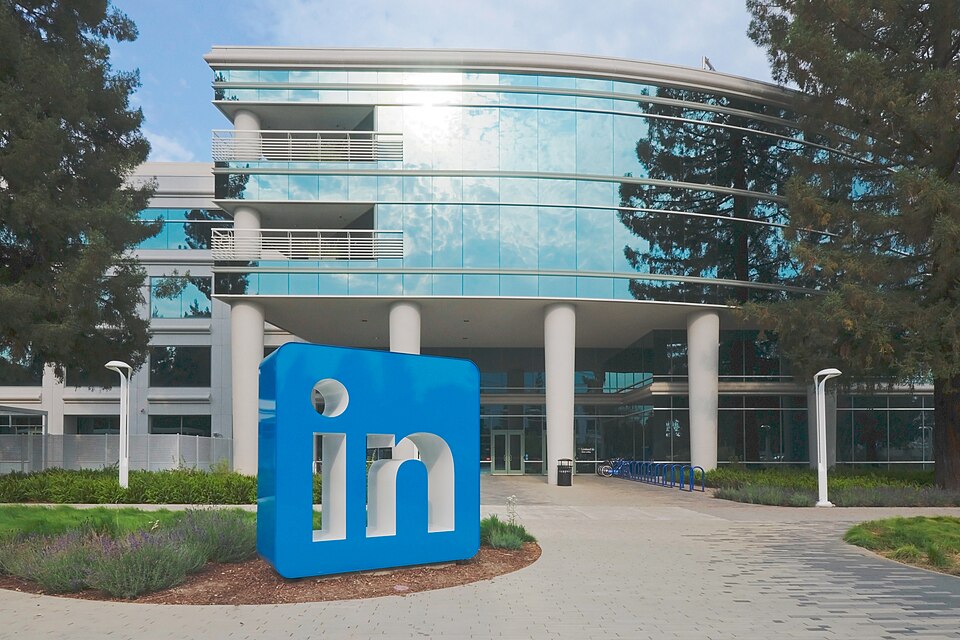The curved glass front of LinkedIn's Sunnyvale headquarters building, with a large blue 'in' logo sculpture standing on the lawn beside the entrance path.