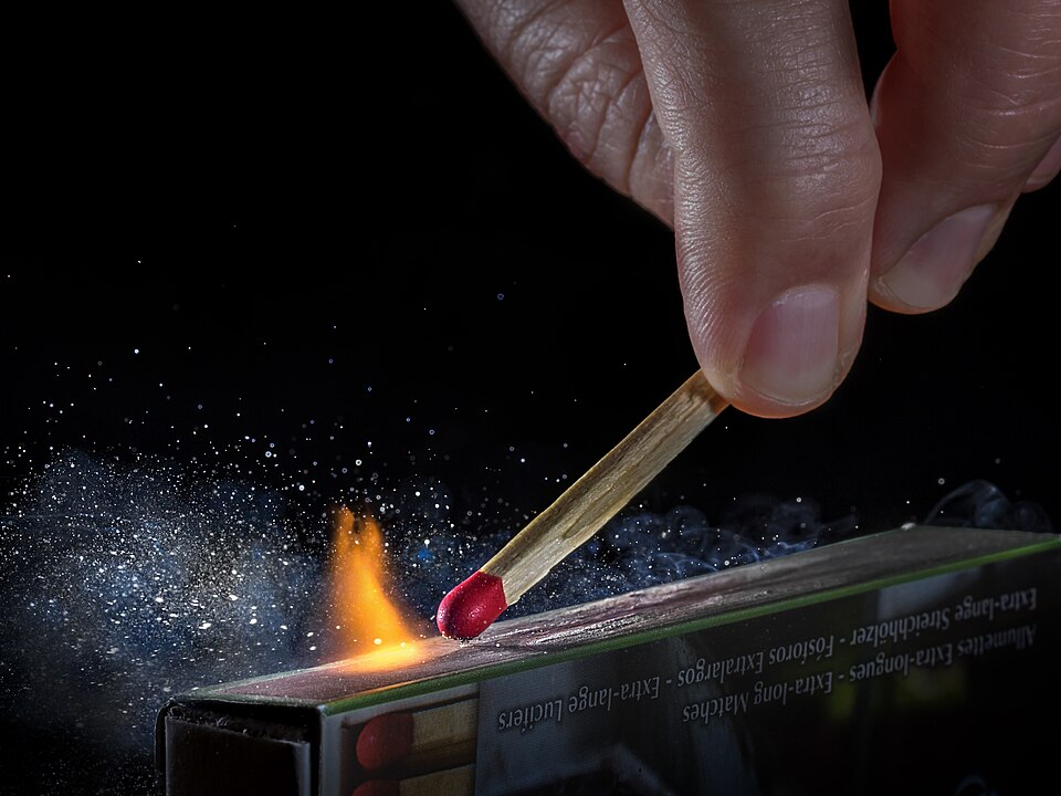 A hand strikes a red-headed safety match along the striking strip on the side of the matchbox, throwing off sparks as the head ignites.