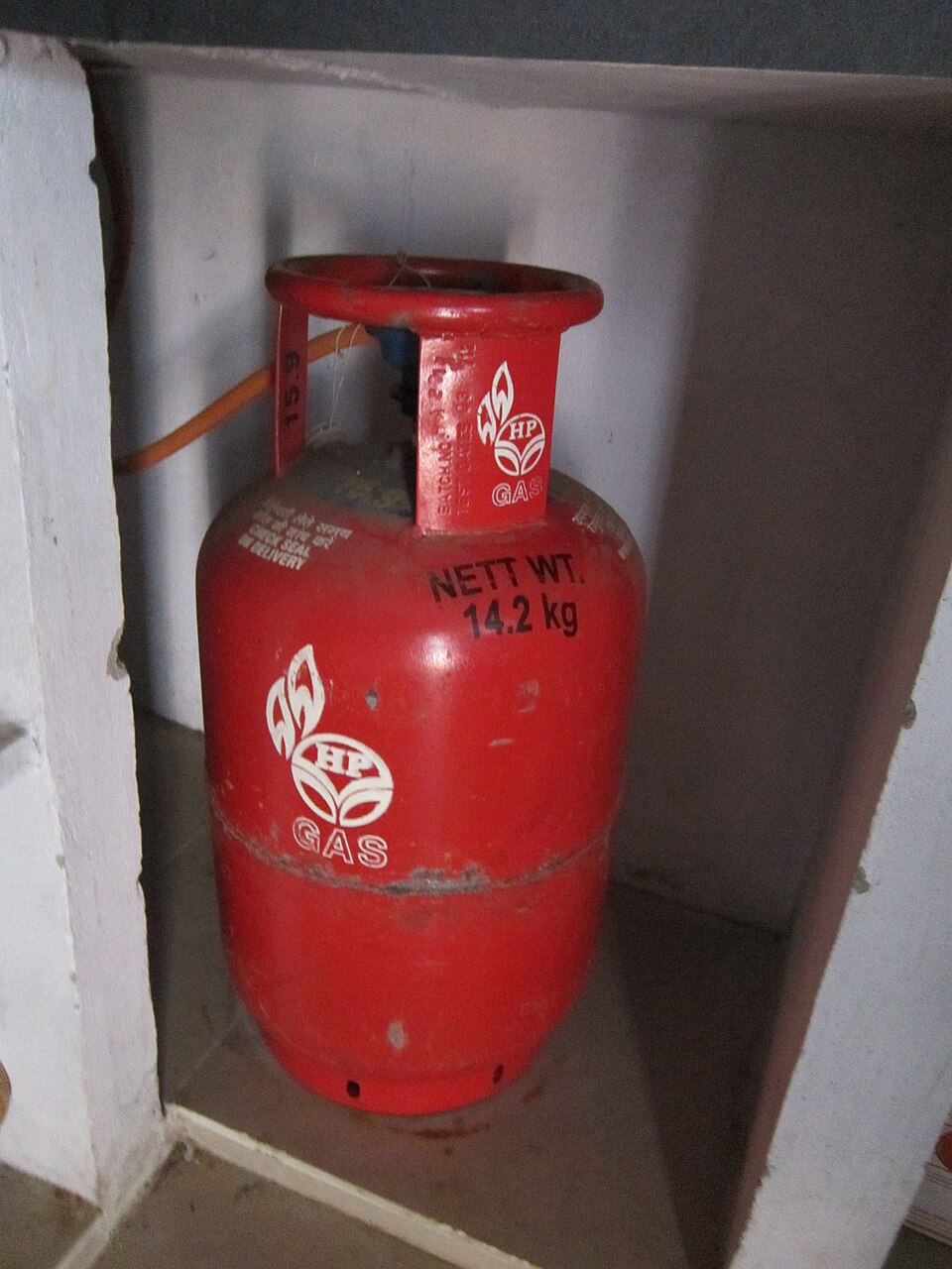 A red Indian domestic LPG cooking gas cylinder marked 'NETT WT. 14.2 kg' standing in a kitchen cabinet with an orange rubber hose attached.