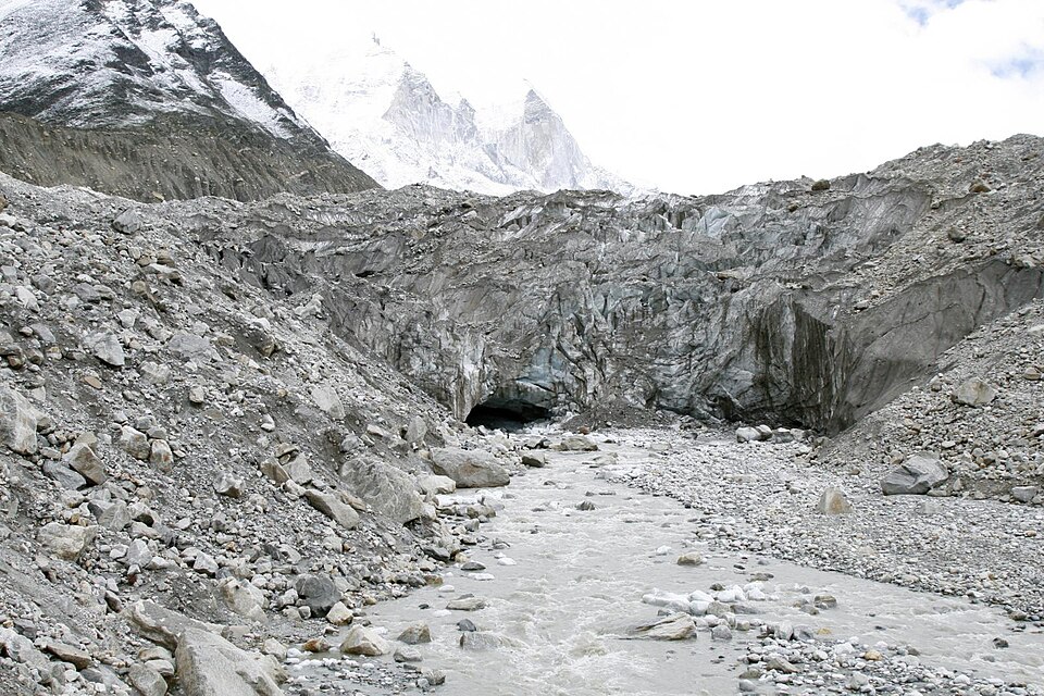 The snout of the Gangotri Glacier at Gaumukh, where a meltwater stream emerges from an ice cave below debris-covered ice and snow peaks.