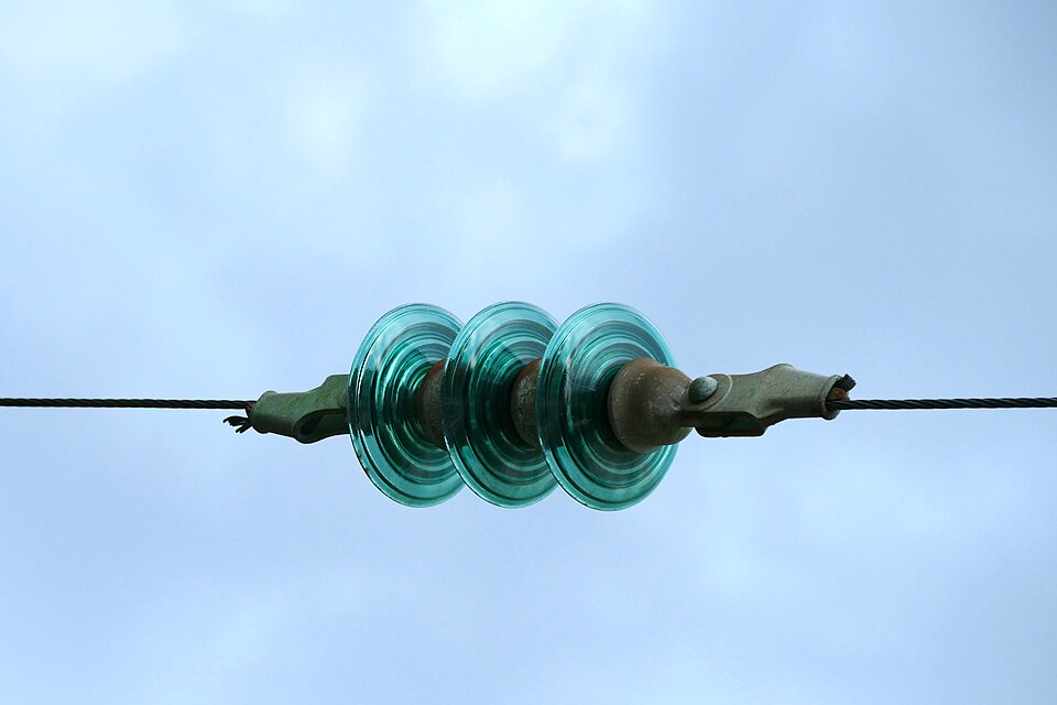 A string of three ribbed glass disc insulators joining two lengths of an overhead power conductor against a pale sky.