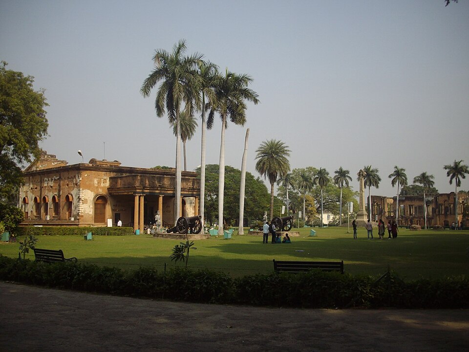 The shell-scarred ruins of the British Residency at Lucknow standing on a lawn, with old cannons displayed in front and palm trees around the site.