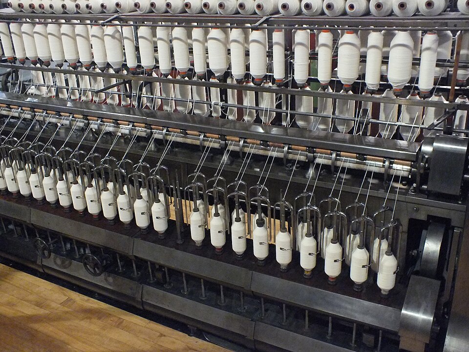 A ring spinning frame in a cotton mill, with rows of white yarn bobbins and cones being wound on the machine's spindles.