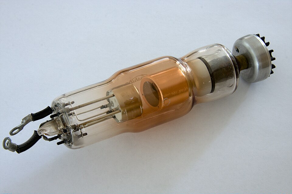 An X-ray tube lying on a white surface, showing the evacuated glass envelope with the filament and cathode assembly at one end and the copper anode block with its target in the middle.