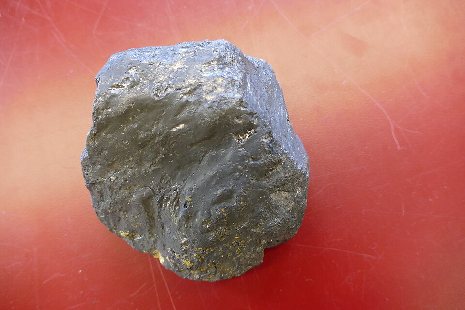 A rough lump of natural graphite with a dull grey metallic sheen, resting on a red surface.