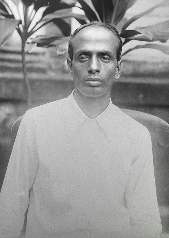 A black-and-white photographic portrait of the revolutionary Surya Sen, known as Masterda, seated in a plain white kurta before a leafy background.