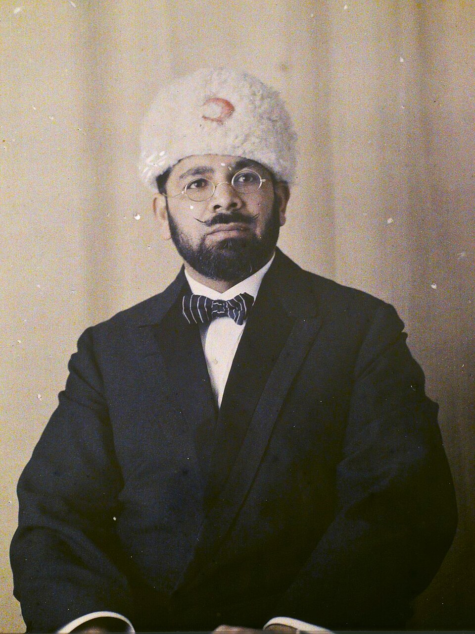 A 1920 colour portrait of Maulana Mohammad Ali Jauhar in a dark suit and bow tie, wearing a white fur cap bearing a crescent.