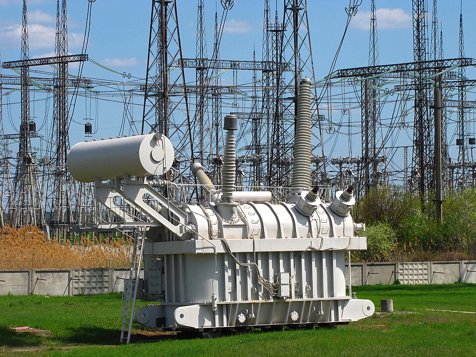 A large white oil-filled high-voltage power transformer with tall bushings standing on a grassy plot inside an outdoor electrical substation, with lattice towers and overhead lines behind it.