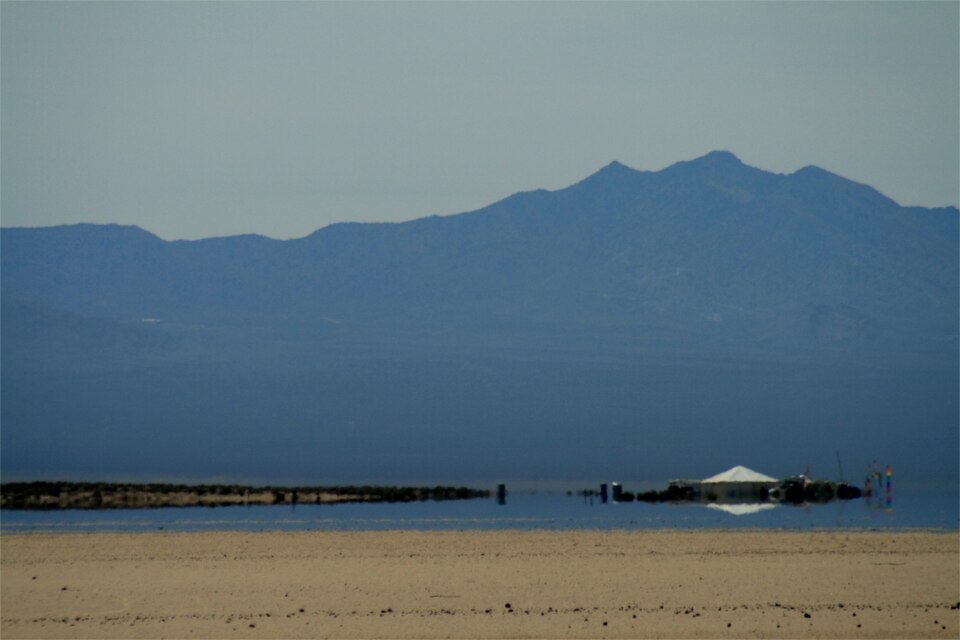 A mirage on a hot desert flat: beyond the dry sandy foreground a sheet of apparent water seems to lie across the plain, reflecting a distant hut and shoreline, with bare mountains behind.