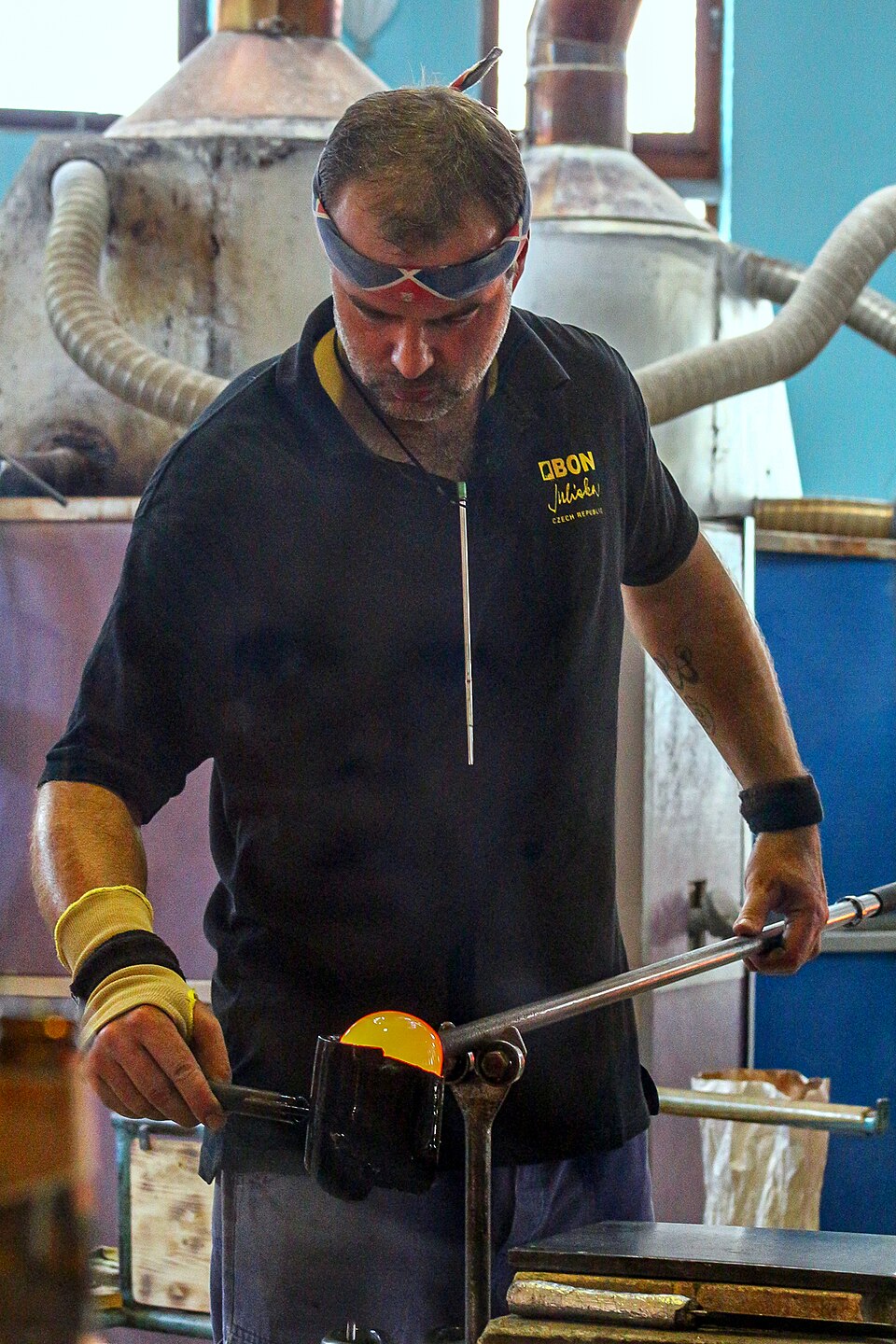 A glassworker in a hot shop rolling a glowing orange blob of molten glass on the end of a long steel blowpipe.