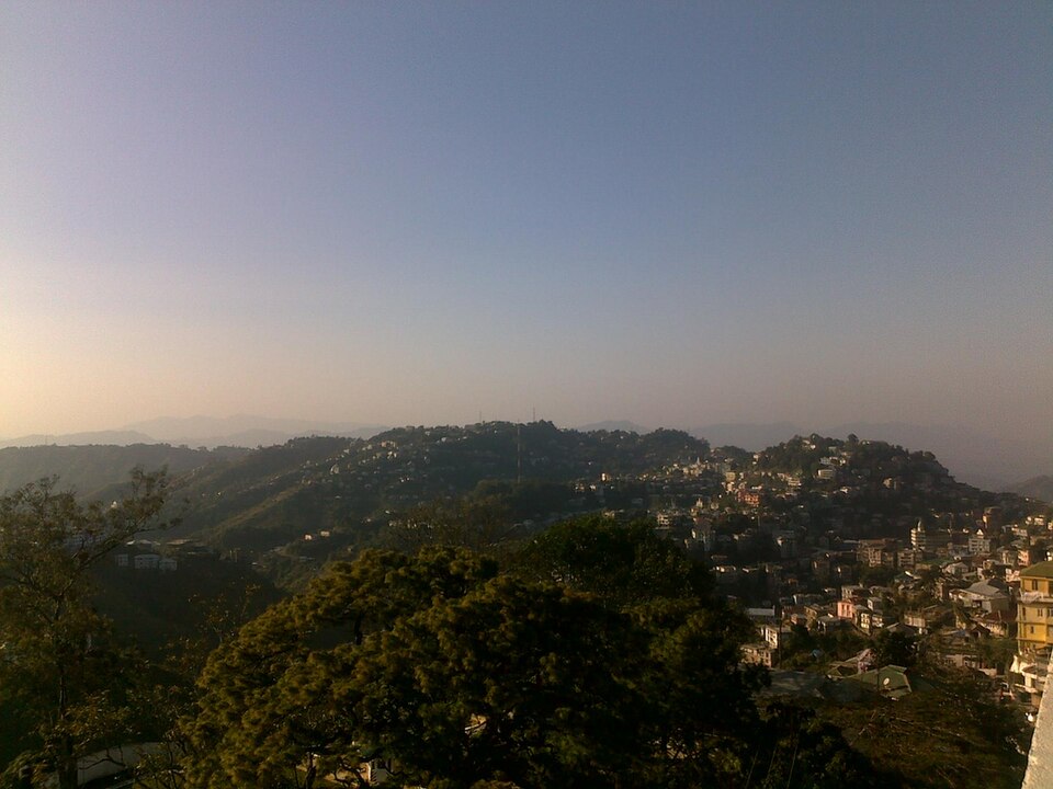 A wide view of Aizawl at dusk, its houses spread along the crests of forested ridges with hazy blue hills receding behind.