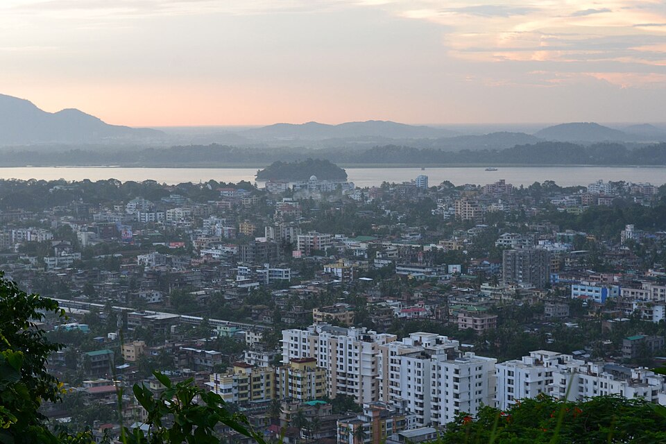 A hilltop view over the dense cityscape of Guwahati at dusk, with the wide Brahmaputra river and its wooded midstream island in the background.