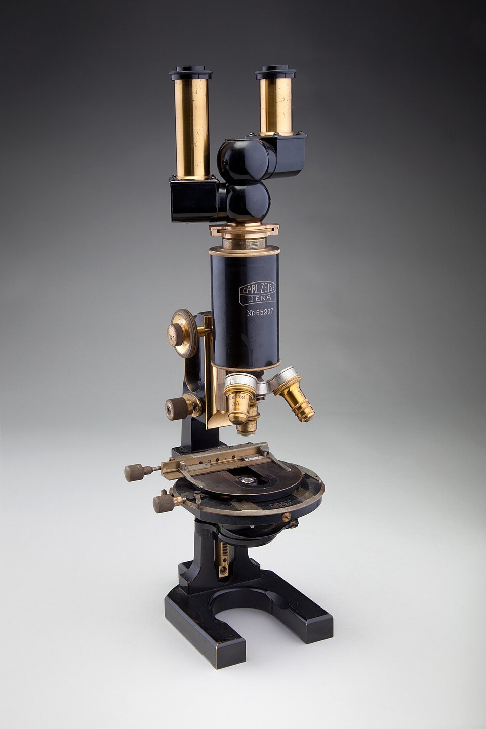 A binocular compound optical microscope on a stand, showing the two eyepiece tubes at the top, the rotating turret of objective lenses above the specimen stage, and the focusing knobs.