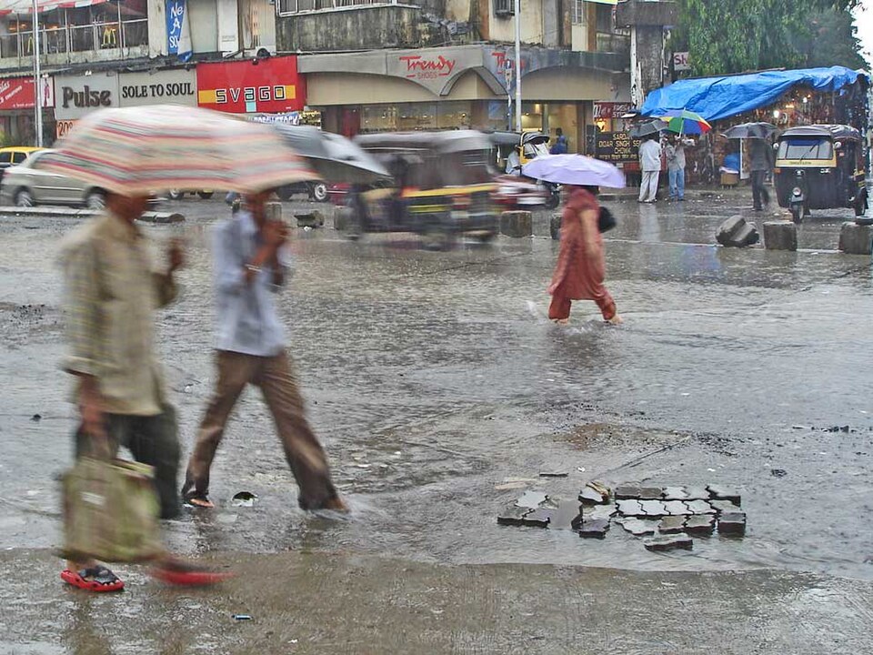 Heavy monsoon rain falling on a waterlogged Mumbai street, with pedestrians crossing under umbrellas and autorickshaws waiting by shopfronts.