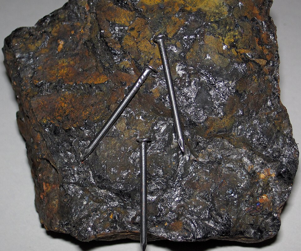 A dark metallic block of magnetite (lodestone) with three iron nails clinging to its surface, held there by the rock's own magnetism.