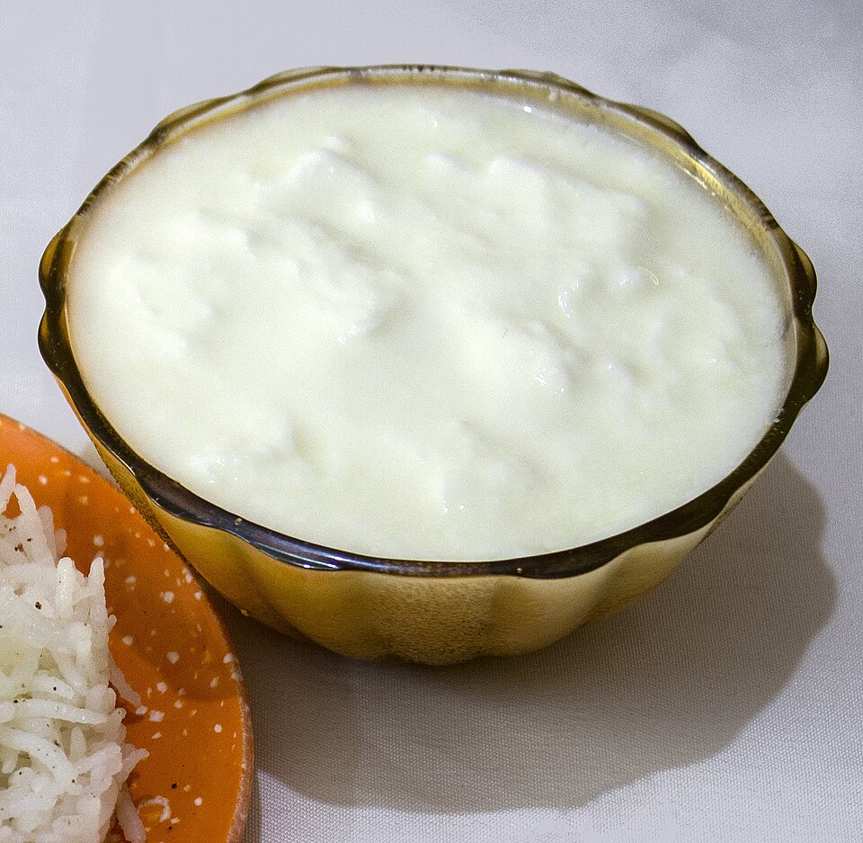 A brass bowl filled to the brim with thick white set curd (dahi), with a plate of plain rice beside it.
