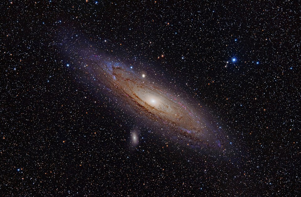 The Andromeda Galaxy seen tilted against a dense star field, its bright core surrounded by dusty spiral arms and two small companion galaxies.