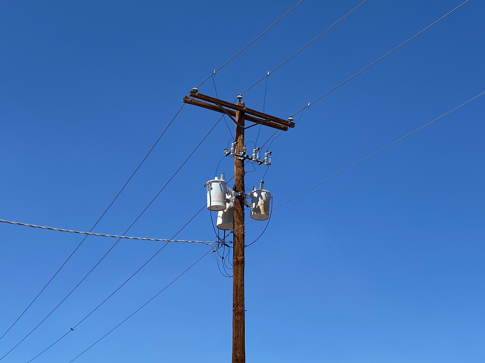Two grey cylindrical distribution transformers mounted on a wooden electricity pole with crossarm and overhead lines against a clear blue sky.