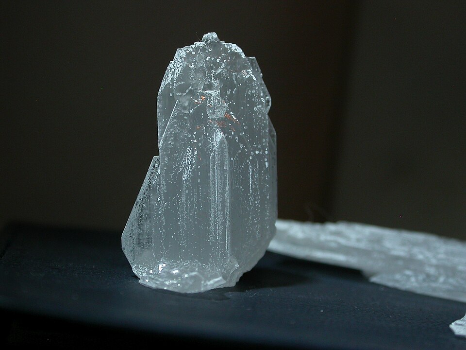 A single translucent, colourless crystal of washing soda (sodium carbonate decahydrate) standing on a dark surface, with a smaller crystal shard beside it.