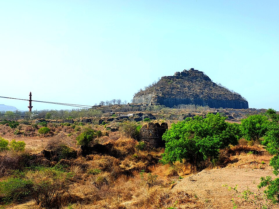 A distant view of Daulatabad (Devagiri) Fort in Maharashtra: a steep conical rock hill with a scarped cliff face and a citadel on its summit, with the tall Chand Minar tower and outer bastions on the scrubby plain below.