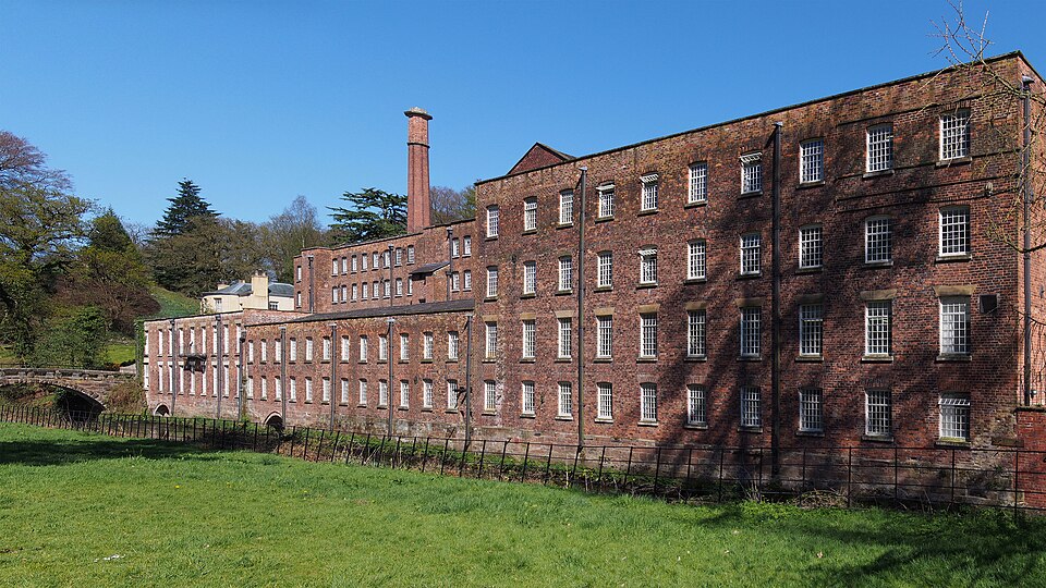 The long red-brick multi-storey cotton mill at Quarry Bank, Styal, with its tall brick chimney, seen across a grassy river bank on a clear day.