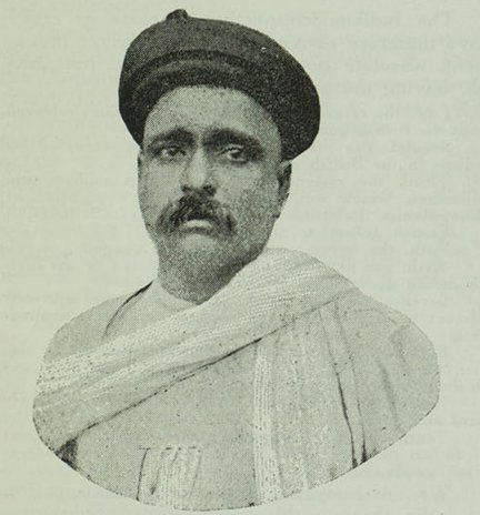An old black-and-white bust portrait of Bal Gangadhar Tilak wearing a Maratha turban and a white shawl, with a heavy moustache.