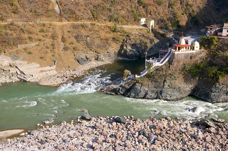 Two Himalayan rivers meeting in a rocky gorge at Rudraprayag, with a small temple and bathing steps on the promontory between them and a boulder bank in the foreground.