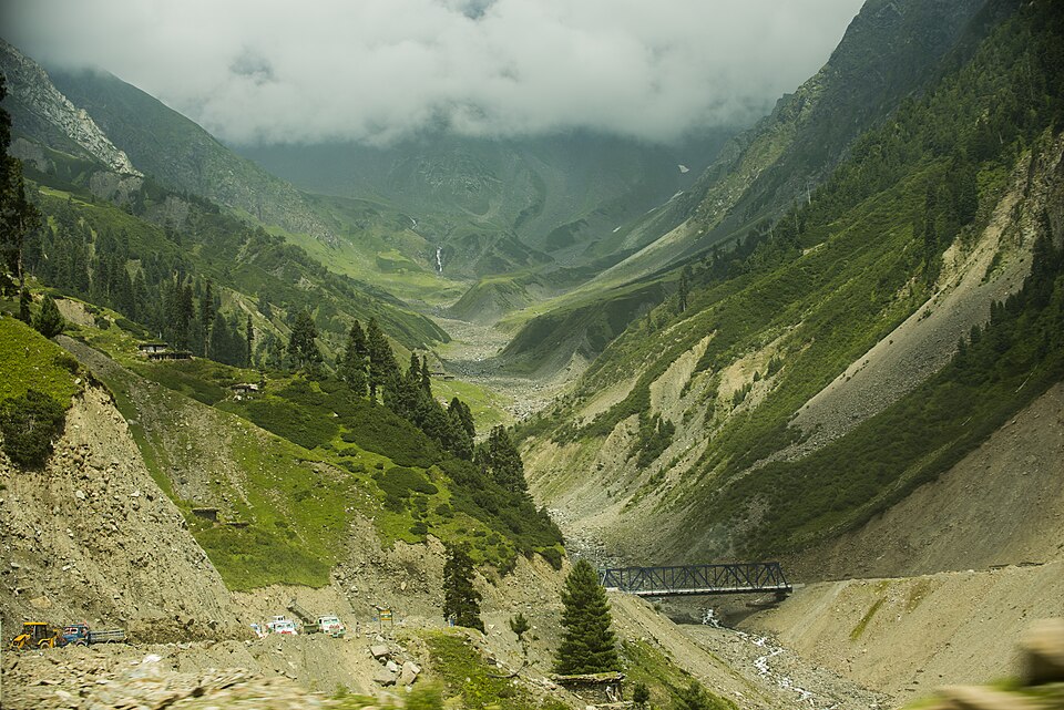 A steep green valley in the Pir Panjal range of the Jammu region at Pir Ki Gali, with conifer-clad slopes, bare scree, a small steel bridge over the stream and low cloud on the ridge above.